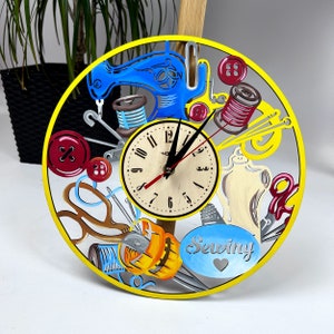 Peut inclure: Une horloge circulaire jaune sur le thème de la couture. Le cadran de l'horloge a des chiffres romains noirs et une aiguille rouge et noire. L'horloge est entourée de divers outils de couture, notamment des ciseaux, des bobines de fil, des boutons et une machine à coudre.