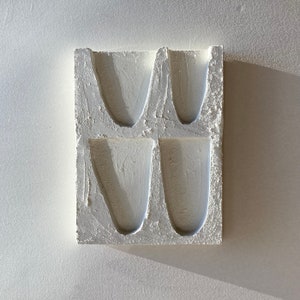 May include: A rectangular, off-white plaster mold with four concave, vase-shaped depressions. The surface has a textured, rough appearance, and the shadows suggest a strong light source from the upper left.