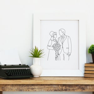 Pode incluir: Uma impress&atilde;o emoldurada branca com um desenho de linha preta de um casal segurando um buqu&ecirc; de flores. A impress&atilde;o est&aacute; em uma mesa de madeira com uma m&aacute;quina de escrever, um vaso com uma planta e uma pilha de livros.