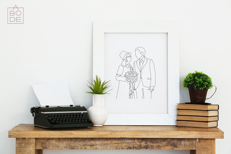Pode incluir: Uma impress&atilde;o emoldurada branca com um desenho de linha preta de um casal segurando um buqu&ecirc; de flores. A impress&atilde;o est&aacute; em uma mesa de madeira com uma m&aacute;quina de escrever, um vaso com uma planta e uma pilha de livros.