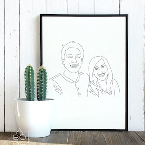 Pode incluir: Uma impress&atilde;o emoldurada preta de um desenho de linha de um casal. O casal est&aacute; de frente para o espectador e sorrindo. A impress&atilde;o est&aacute; em um fundo branco e est&aacute; sentada em uma superf&iacute;cie de madeira.