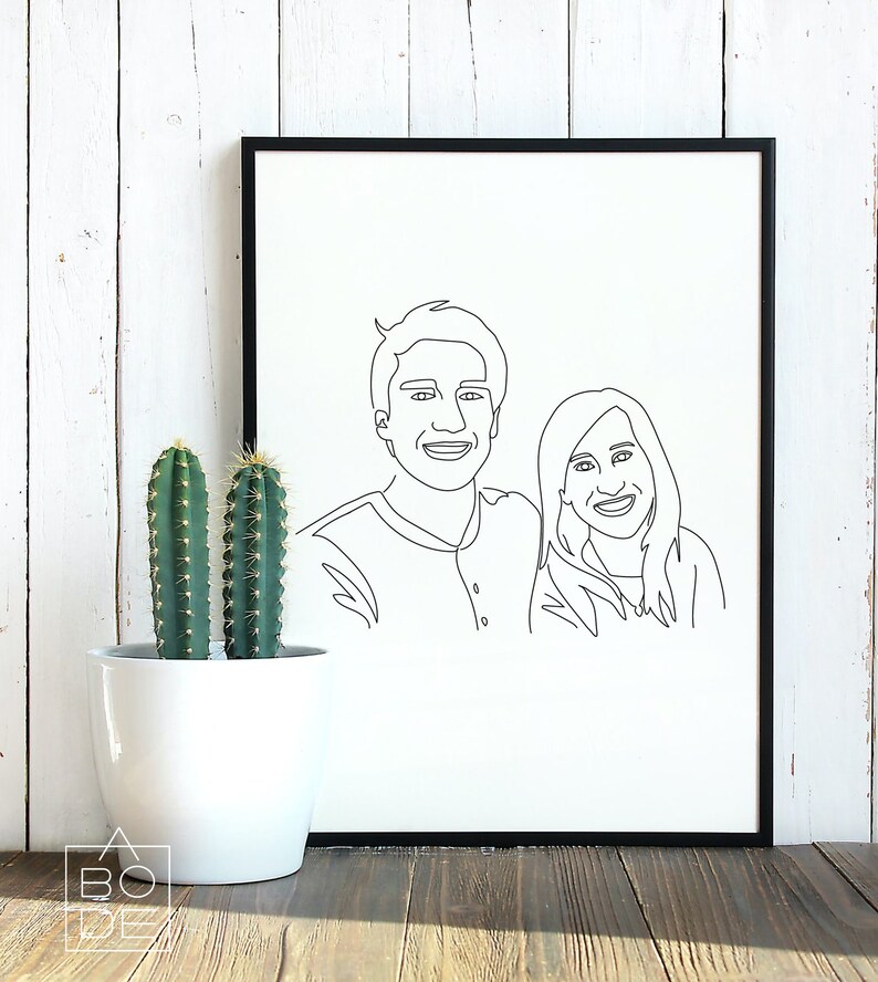 Pode incluir: Uma impress&atilde;o emoldurada preta de um desenho de linha de um casal. O casal est&aacute; de frente para o espectador e sorrindo. A impress&atilde;o est&aacute; em um fundo branco e est&aacute; sentada em uma superf&iacute;cie de madeira.