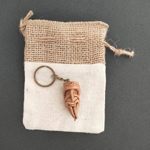 Pode incluir: Um chaveiro de couro feito à mão com um design de rosto estilizado, preso a um porta-chaves de metal. O chaveiro é exibido em uma bolsa de tecido branco com uma parte superior de serapilheira, amarrada com um cordão de barbante natural. O fundo é cinza escuro.