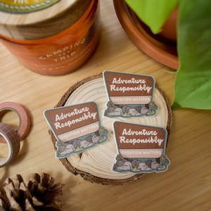 May include: Three brown and white stickers with the text "Adventure Responsibly, Creation Matters" on a wooden surface. The stickers feature a cartoon illustration of a group of animals.