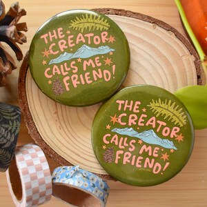 May include: Two olive green round buttons with the text "THE CREATOR CALLS ME FRIEND!" in pink and white. The buttons are on a wooden slice, with decorative tapes and a pine cone nearby. The buttons have a mountain and star design.