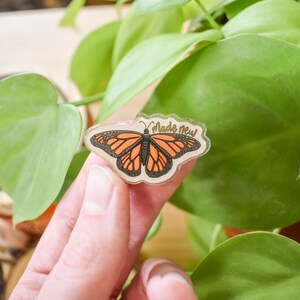 May include: A clear, butterfly-shaped sticker with orange and black wings and the text "Made New." The sticker is held in front of a green plant. The butterfly design resembles a monarch butterfly.