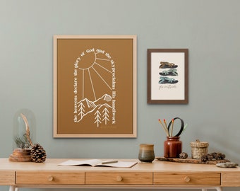 Op de afbeelding: Een bruin ingelijst print met een wit berg- en zonontwerp. De tekst luidt "the heavens declare the glory of God and the sky proclaims His handiwork". Een kleiner bruin ingelijst print met een witte achtergrond en de tekst "go outside" bevindt zich boven een houten bureau met een glazen stolp, een dennenappel, een bruine keramische mok, een houten dienblad met een bruine pot met potloden en een geweven mand.