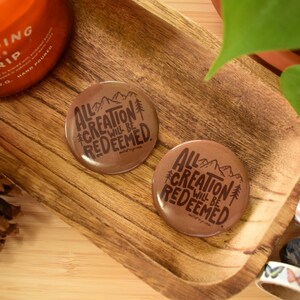 May include: Two brown circular buttons with the text "All Creation Will Be Redeemed" and mountain and tree graphics. The buttons are on a wooden tray with decorative tape rolls, a pine cone, and an orange candle.