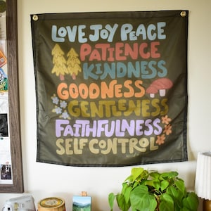 May include: A black wall hanging with the words "Love Joy Peace Patience Kindness Goodness Gentleness Faithfulness Self Control" in colorful, retro-style lettering. The design includes illustrations of trees and a mushroom.