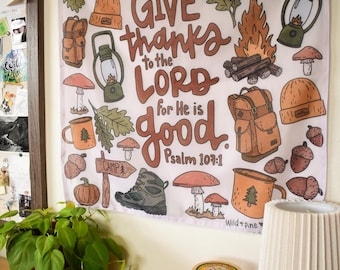 Op de afbeelding: Een witte wandhanger met een campingthema. Het bevat illustraties van pompoenen, eikels, paddenstoelen, een lantaarn en een rugzak. De tekst "GIVE thanks to the LORD for He is good Psalm 107:1" staat prominent afgebeeld.