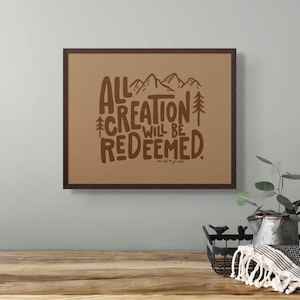 Puede incluir: Una impresión enmarcada en marrón con un fondo beige que presenta una ilustración dibujada a mano de montañas y árboles con el texto "All Creation Will Be Redeemed." El texto está en marrón y la firma del artista "wild+pine" está en marrón más pequeño en la parte inferior.