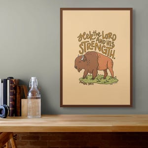 Puede incluir: Una impresión enmarcada con un fondo beige que presenta un bisonte marrón y naranja con el texto "Seek the Lord and His Strength." El texto está en marrón y dorado. La impresión está enmarcada en un marco marrón.