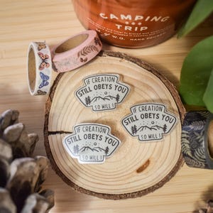 May include: Three white and black enamel pins with the text "If creation still obeys you, so will I."  The pins are on a wooden surface with a brown background.