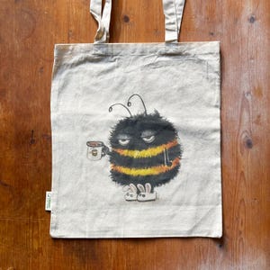 May include: A white tote bag with a black and yellow bee illustration wearing bunny slippers and holding a cup of coffee. The bee has a sleepy expression and is wearing a black and yellow striped body with fuzzy black fur. The bag is made of organic cotton.