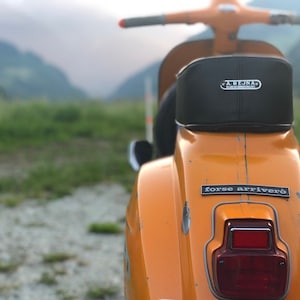 Könnte beinhalten: Nahaufnahme eines orangefarbenen Vintage-Rollers mit schwarzem Sitz und dem Text "FORSE ARRIVERÒ". Der Roller hat ein rotes Rücklicht. Im Hintergrund Berge und ein Schotterboden.