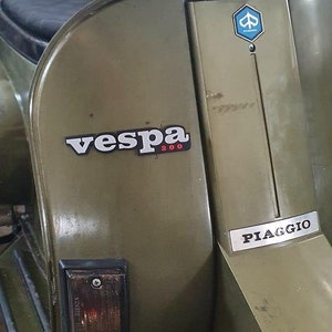 Può includere: Uno scooter Vespa 200 verde con un logo Vespa argentato e un logo Piaggio argentato. Lo scooter ha una sella nera e un faro nero.