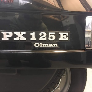 Può includere: Primo piano di uno scooter nero con un badge argentato PX125E e il testo Olman. Lo scooter ha una luce posteriore cromata e uno pneumatico nero.