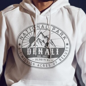 May include: White hooded sweatshirt with a gray print of a national park logo. The logo features a mountain range, a sun, and the text "National Park", "Denali", "est. 1917", "6.1 Million Acres of Beauty", and "20,310 ft EL".