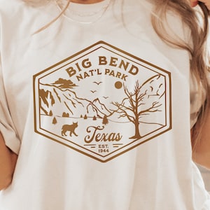 Könnte beinhalten: Ein weißes T-Shirt mit einer braunen Grafik eines Vintage-Logos für den Big Bend National Park in Texas. Das Logo zeigt eine Bergkette, einen Baum, eine Sonne und einen Kojoten.