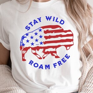 Könnte beinhalten: Weißes T-Shirt mit einem blauen und roten amerikanischen Flaggen-Design in Form eines Büffels. Der Text "Stay Wild Roam Free" ist um den Büffel herum geschrieben.