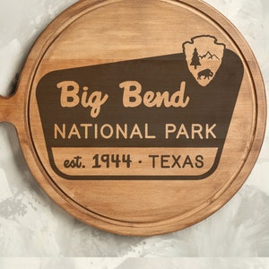 May include: A round wooden cutting board with a handle. The board is engraved with the words "Big Bend National Park est. 1944 - Texas" and a stylized image of a mountain range and a bear.