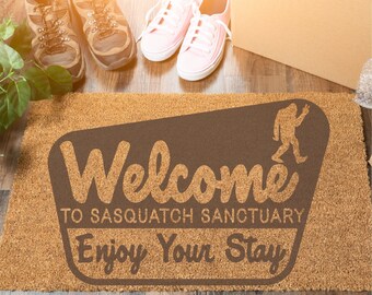 May include: A brown coir doormat with the text "Welcome to Sasquatch Sanctuary Enjoy Your Stay" in a brown rectangle with a stylized Sasquatch figure walking to the right.