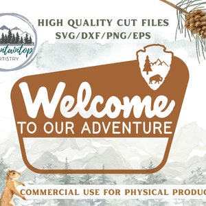 May include: A brown sign with rounded corners and a white border reads "Welcome to our adventure". The sign is set against a watercolor background of mountains and trees. The text "Mountaintop Artistry" is in a circle with a mountain scene in the background.