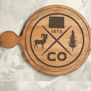 May include: A brown wooden serving board with a handle. The board has a design of a mountain range, a bighorn sheep, a pine tree, and the text "CO 1876".