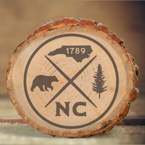 Może przedstawiać: Okrągły drewniany znak z brązowym niedźwiedziem, sosną, stanem Karolina Północna i rokiem 1789. Tekst "NC" znajduje się w środku znaku.