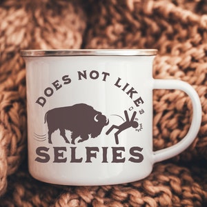 May include: White enamel mug with a brown silhouette of a buffalo and a stick figure running away from it. The text on the mug reads "Does not like selfies".