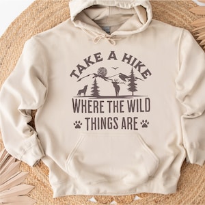 May include: A beige hoodie with a brown graphic print that says "Take a Hike Where the Wild Things Are". The graphic features a mountain range, a deer, a wolf, and trees.