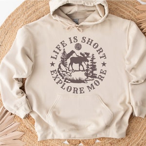 May include: A beige hoodie with a brown graphic print of a moose standing in a forest with the text "Life is Short Explore More" in a circle around the image.