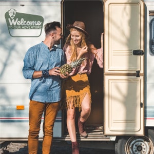 May include: A couple standing in the doorway of a white camper van. The woman is wearing a brown hat, a pink shirt, and a brown skirt with fringe. The man is wearing a blue striped shirt, brown trousers, and a blue bandana. They are holding a pineapple.