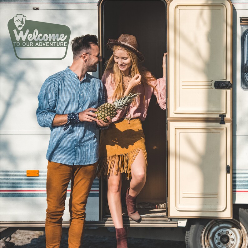 May include: A couple standing in the doorway of a white camper van. The woman is wearing a brown hat, a pink shirt, and a brown skirt with fringe. The man is wearing a blue striped shirt, brown trousers, and a blue bandana. They are holding a pineapple.