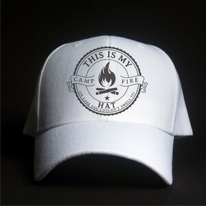 May include: White baseball cap with a curved brim. The cap features a circular graphic with the text "THIS IS MY CAMP FIRE HAT (IN CASE YOU COULDN'T SMELL IT)" surrounding a campfire illustration.