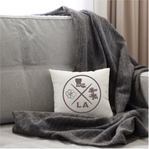 May include: White throw pillow with a brown circle design featuring a magnolia flower, a cypress tree, the outline of Louisiana, and the text "1812" and "LA".