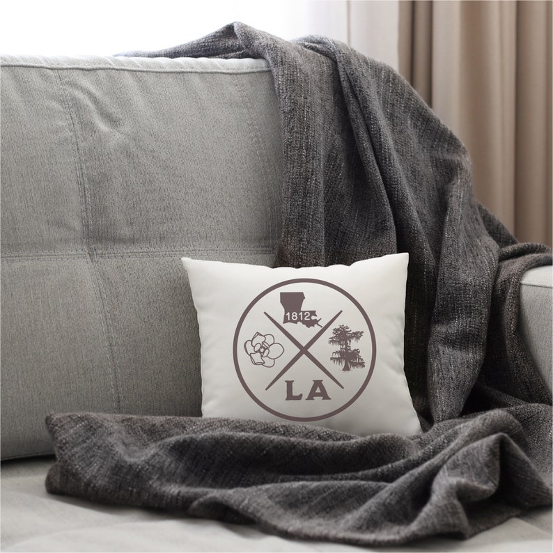 May include: White throw pillow with a brown circle design featuring a magnolia flower, a cypress tree, the outline of Louisiana, and the text "1812" and "LA".