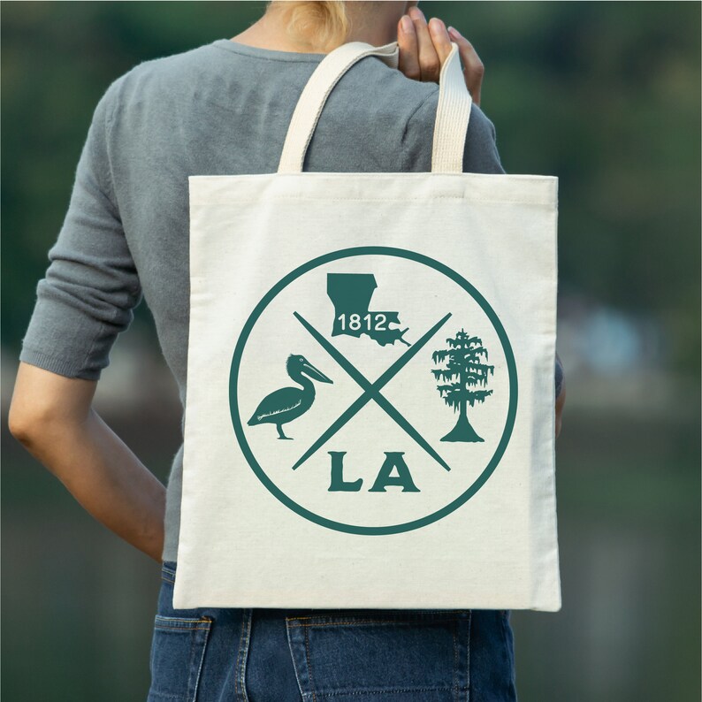 May include: A white canvas tote bag with a green circle design featuring a pelican, a cypress tree, the outline of Louisiana, and the text "LA" and "1812".