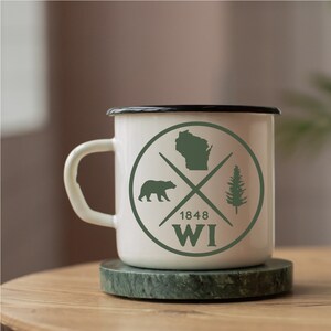 Pode incluir: Caneca de esmalte branco com borda preta e um design circular verde. O c&iacute;rculo apresenta uma silhueta do estado de Wisconsin, um urso, um pinheiro e o texto "1848 WI". A caneca est&aacute; sobre um descanso de m&aacute;rmore verde.