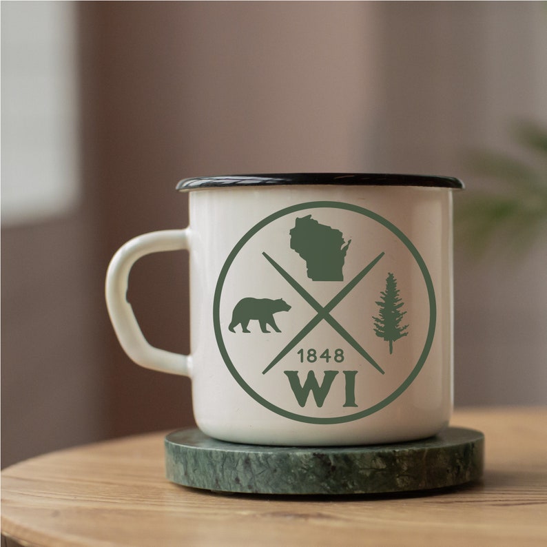 Pode incluir: Caneca de esmalte branco com borda preta e um design circular verde. O c&iacute;rculo apresenta uma silhueta do estado de Wisconsin, um urso, um pinheiro e o texto "1848 WI". A caneca est&aacute; sobre um descanso de m&aacute;rmore verde.
