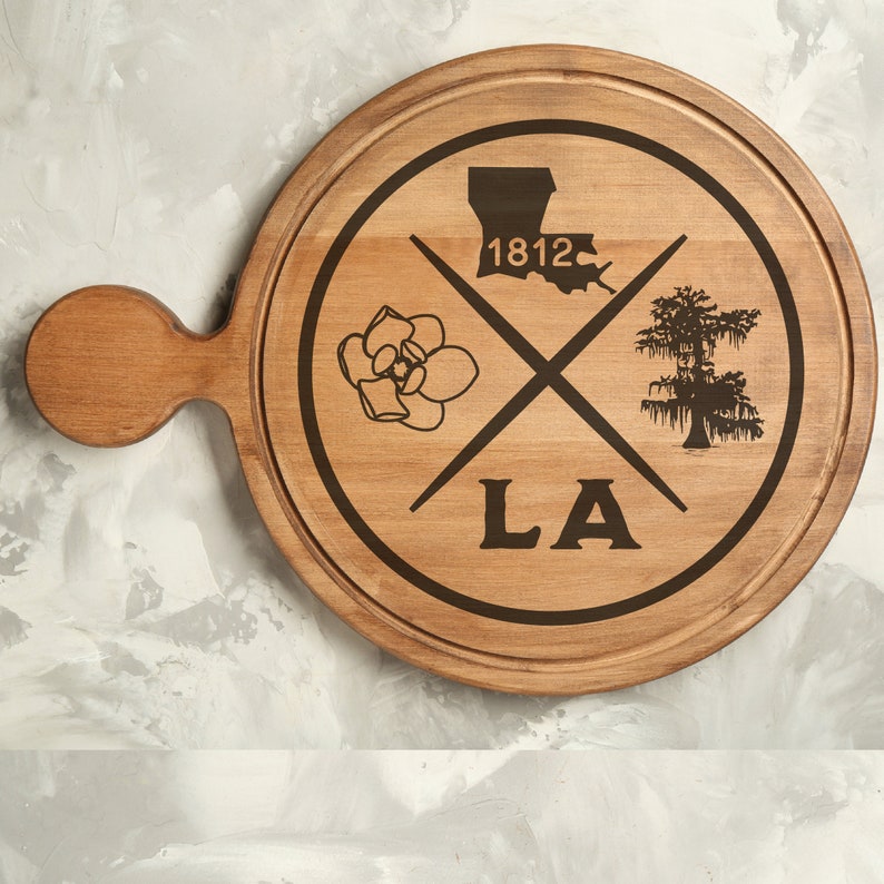 May include: A wooden serving board with a handle, featuring a design of a magnolia flower, a cypress tree, and the outline of Louisiana with the year 1812 and the letters LA.