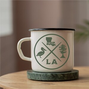May include: White enamel mug with a black rim and a green graphic design. The design features a pelican, a tree, the state of Louisiana, and the text "LA" and "1812".