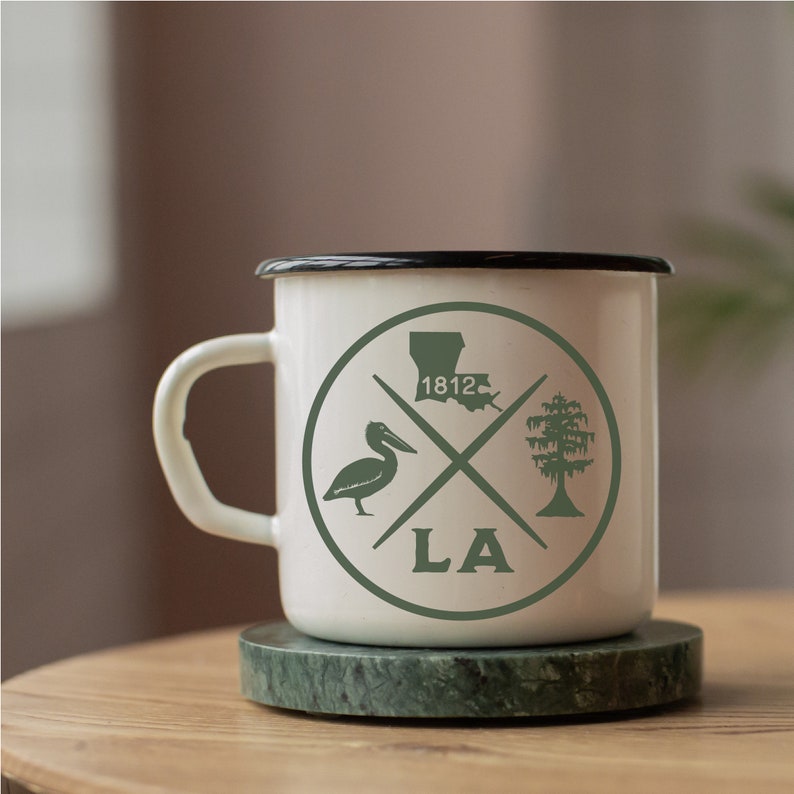 May include: White enamel mug with a black rim and a green graphic design. The design features a pelican, a tree, the state of Louisiana, and the text "LA" and "1812".