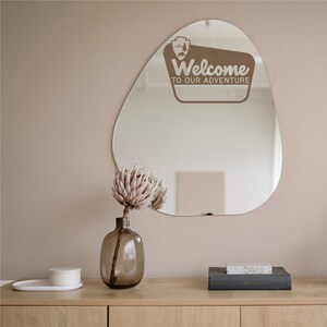 May include: A brown, irregularly shaped mirror with a white sticker that says "Welcome to our adventure". The mirror is hanging on a light pink wall above a wooden dresser with a vase of dried flowers and a small white box.