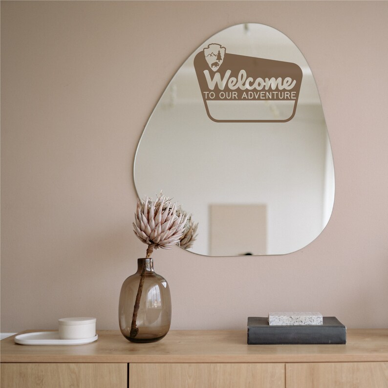 May include: A brown, irregularly shaped mirror with a white sticker that says "Welcome to our adventure". The mirror is hanging on a light pink wall above a wooden dresser with a vase of dried flowers and a small white box.