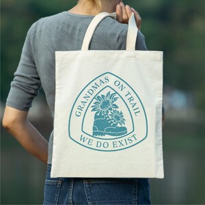 May include: A white tote bag with a blue design featuring a hiking boot with sunflowers and the text "Grandmas on Trail We Do Exist".