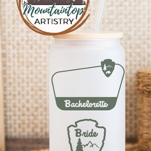 May include: White tumbler with a green mountain and tree design. The tumbler has a bamboo lid and a clear straw. The text "Bride" and "Bachelorette" are printed on the tumbler.
