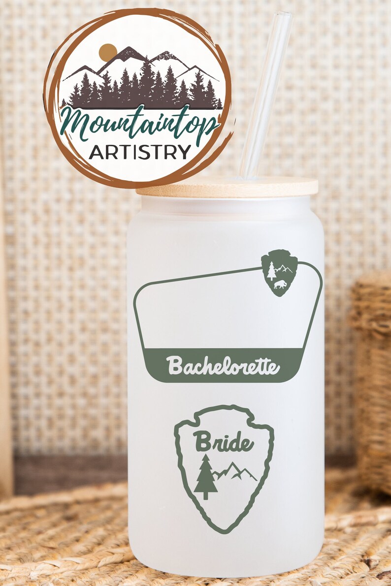 May include: White tumbler with a green mountain and tree design. The tumbler has a bamboo lid and a clear straw. The text "Bride" and "Bachelorette" are printed on the tumbler.
