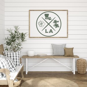 May include: A framed print with a green circle design featuring a magnolia flower, a cypress tree, the state of Louisiana, and the year 1812. The circle is surrounded by the letters "LA".