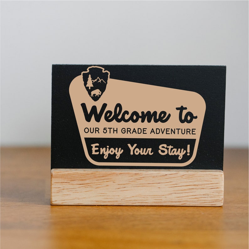 May include: A black chalkboard sign with a wooden base. The sign has a tan background with the text "Welcome to our 5th grade adventure. Enjoy your stay!" and a black silhouette of a park ranger badge with a tree and a buffalo.